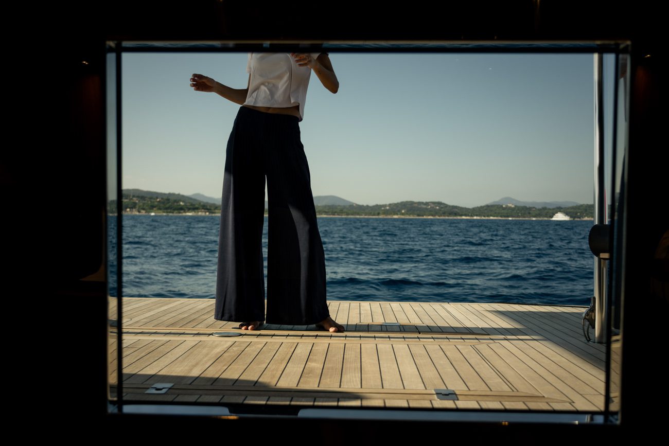 A model standing on the exterior deck of a yacht, framed through an opening with the sea in the background.