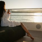 A woman relaxing on the exterior deck of a yacht, overlooking the sea in warm sunset light.
