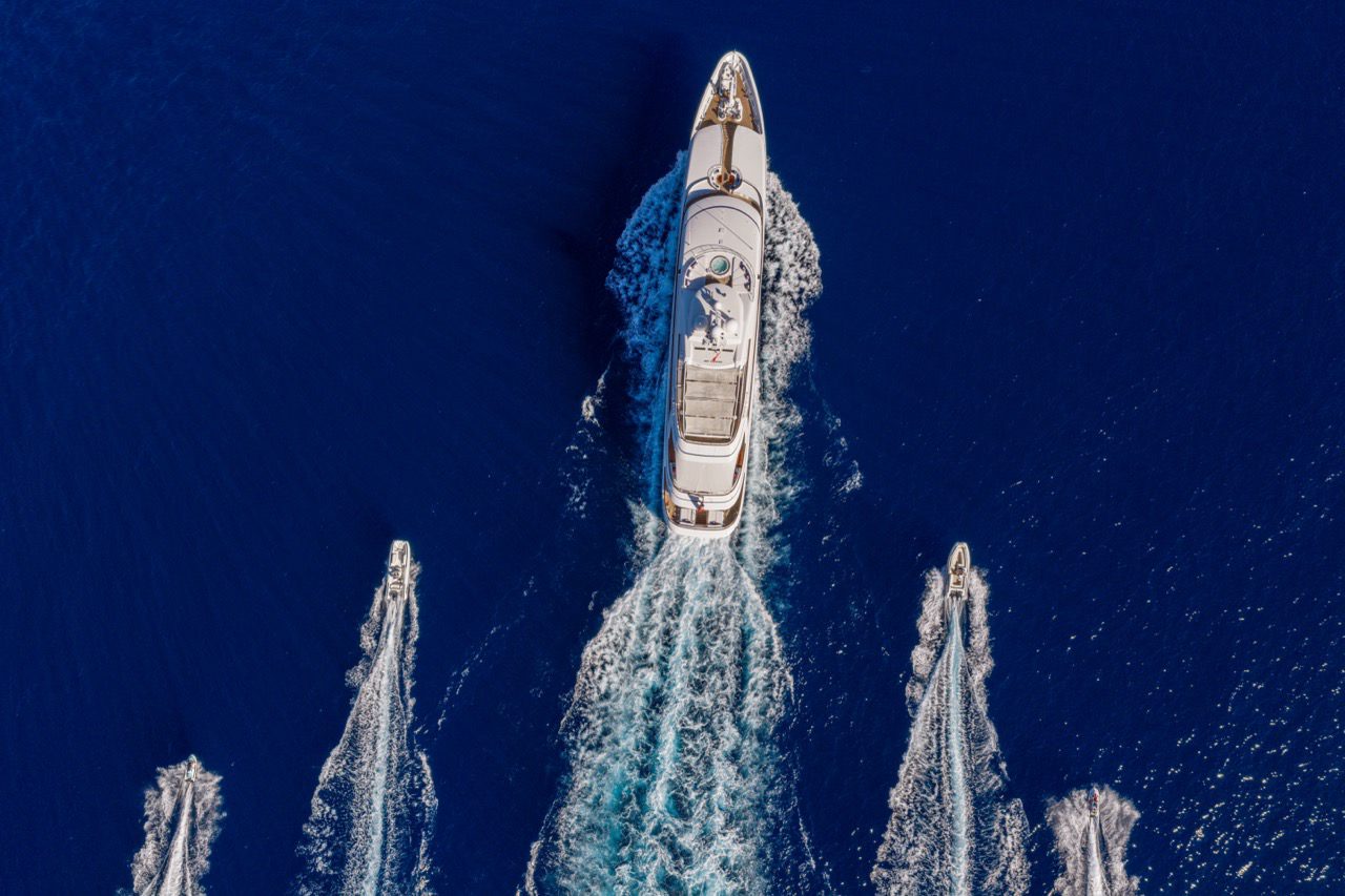 Aerial drone view of a 64-metre Feadship yacht cruising in deep blue water off Palma de Mallorca with two tenders alongside.