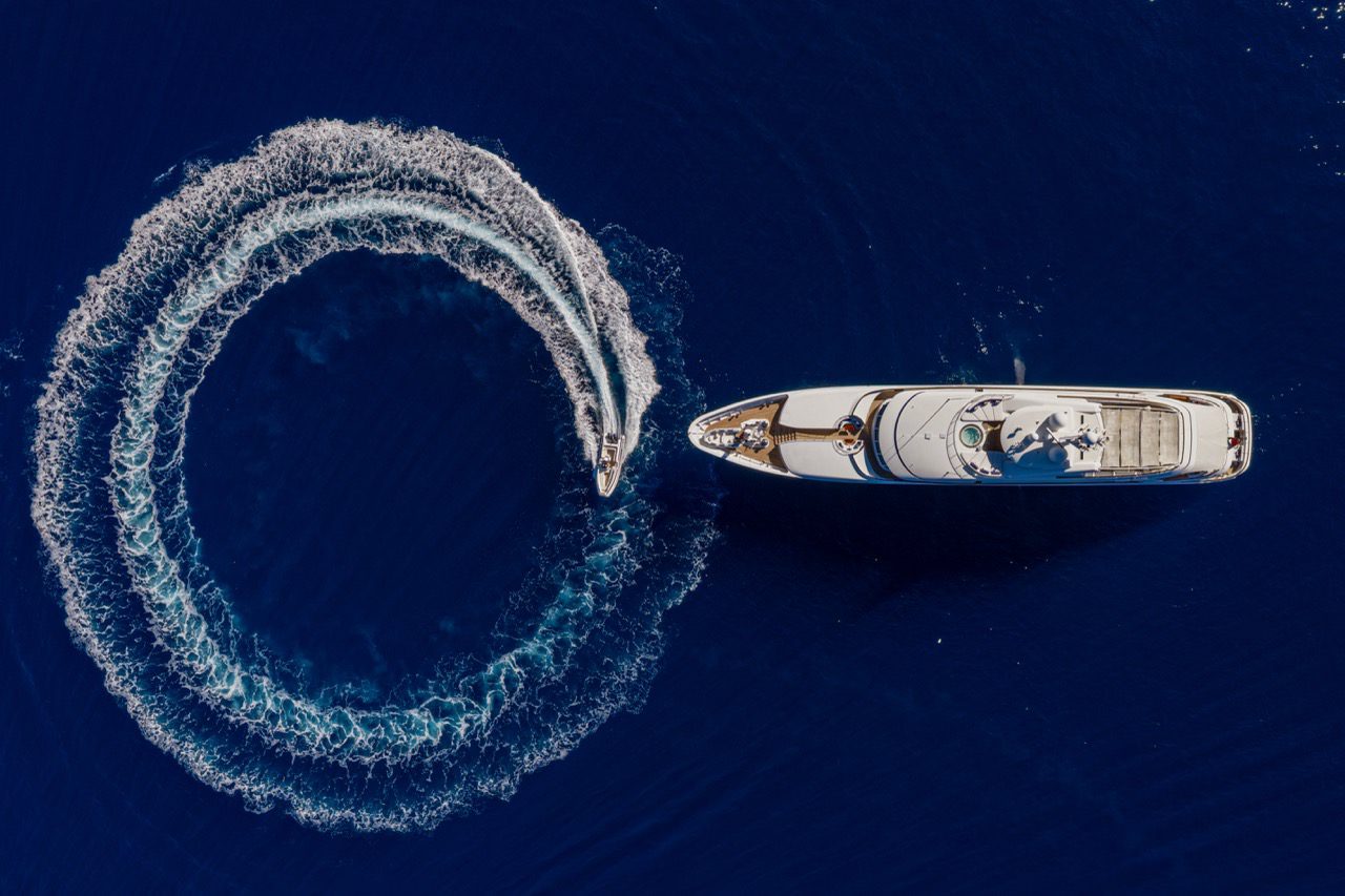 Top-down drone shot of a 64-metre Feadship yacht in Mallorca with a tender circling beside it in clear blue sea.