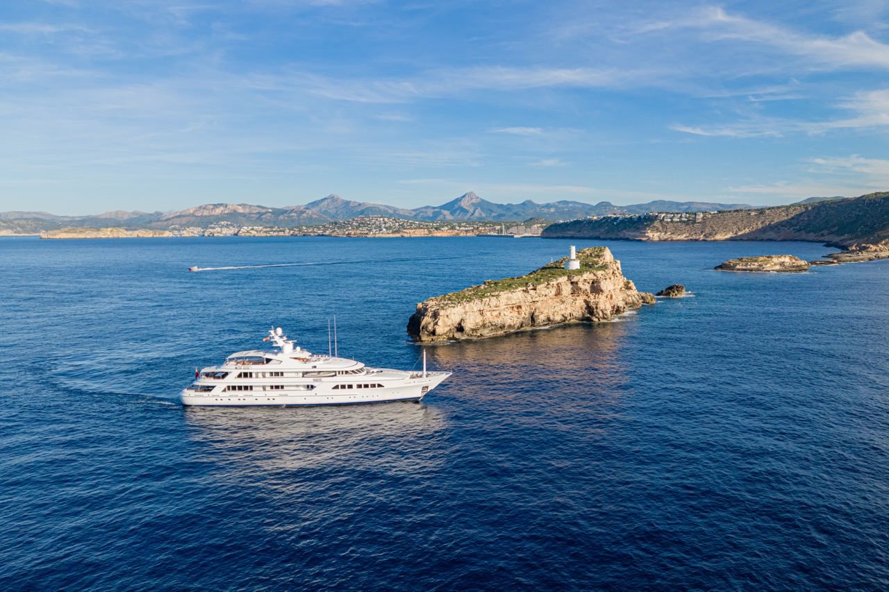 1. Wide aerial view of a 64-metre Feadship yacht sailing off the coast of Palma de Mallorca with rocky shoreline and open sea in the background.